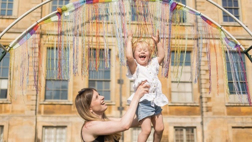 A woman holding up a young child so that their head is touching the multicoloured strings that are falling from the top of a semi-circled piece of metal. Both parent and child have huge grins on their faces.